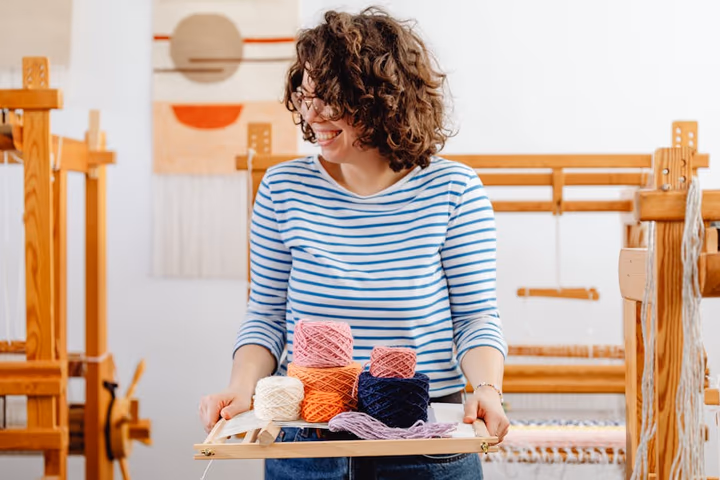 Femme souriante manipulant des fils colorés dans son atelier artisanal, entourée de métiers à tisser.